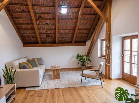 Podkrovní obývací pokoj s dřevěnou podlahou, odkrytými trámy a velkými okny, moderní styl a světlý interiér. | Prodej - dům/vila, 100 m²