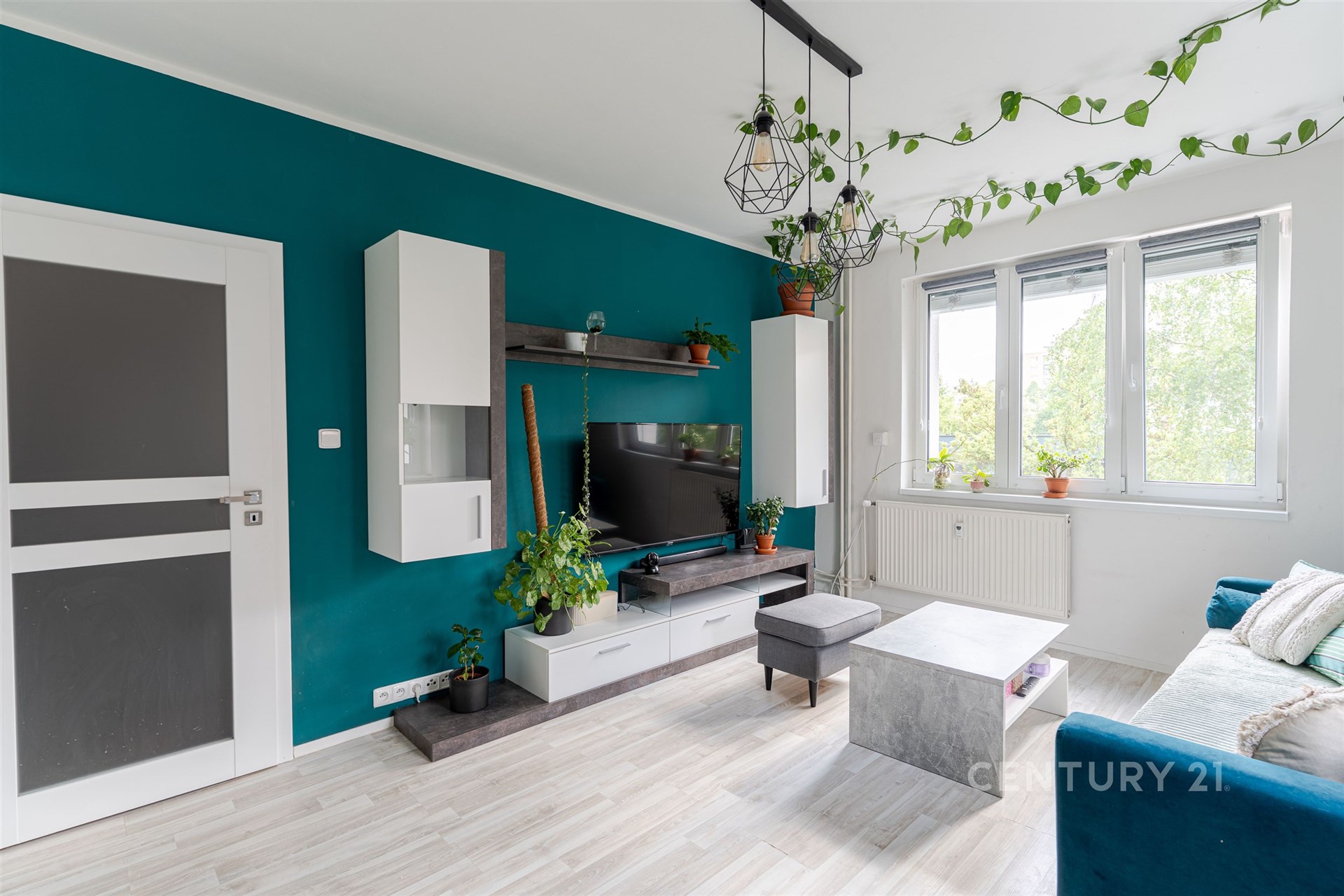 Světlý obývací pokoj s moderními prvky, parketovou podlahou, velká okna a minimalistický design, zdobený rostlinami a stylovým nábytkem.