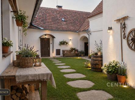 Venkovní dvorek s kamennou dlažbou, světlá fasáda, dekorativní lampy a květiny, rustikální styl, příjemná atmosféra pro relaxaci. | Prodej - dům/vila, 160 m²