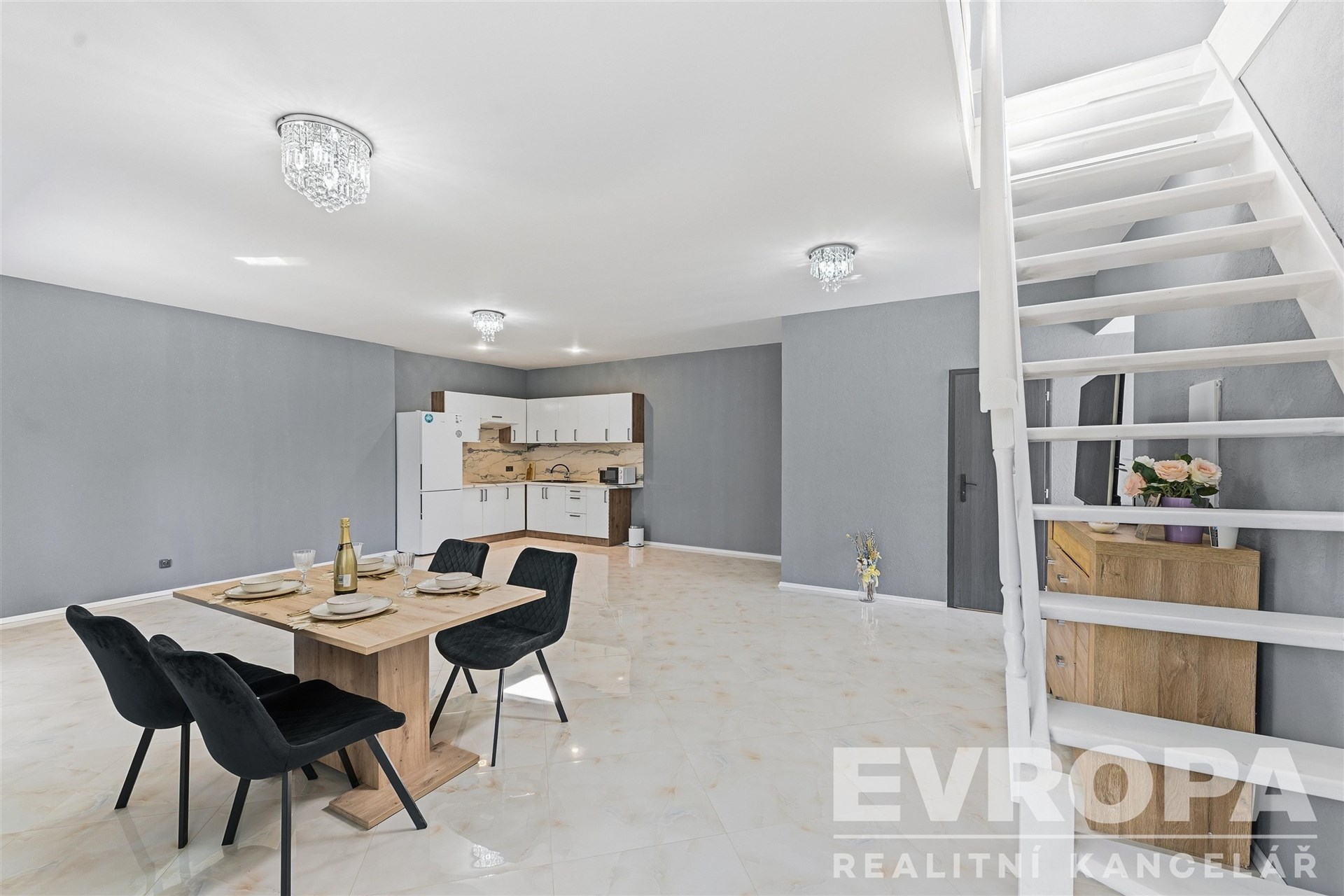 Prostorná jídelna s moderní dlažbou, dřevěným nábytkem a vestavěnou kuchyňskou linkou. Velká okna a elegantní osvětlení dodávají prostoru světlo.