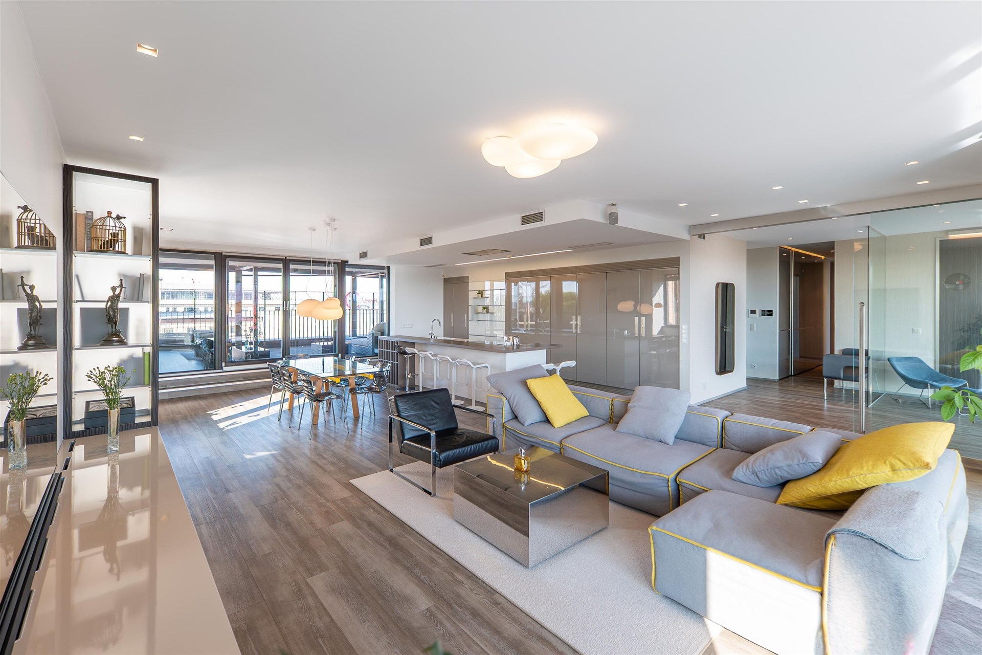 Světlý obývací pokoj s moderní parketovou podlahou, velkými okny a elegantním nábytkem. Otevřený prostor propojený s kuchyní nabízí komfortní bydlení.