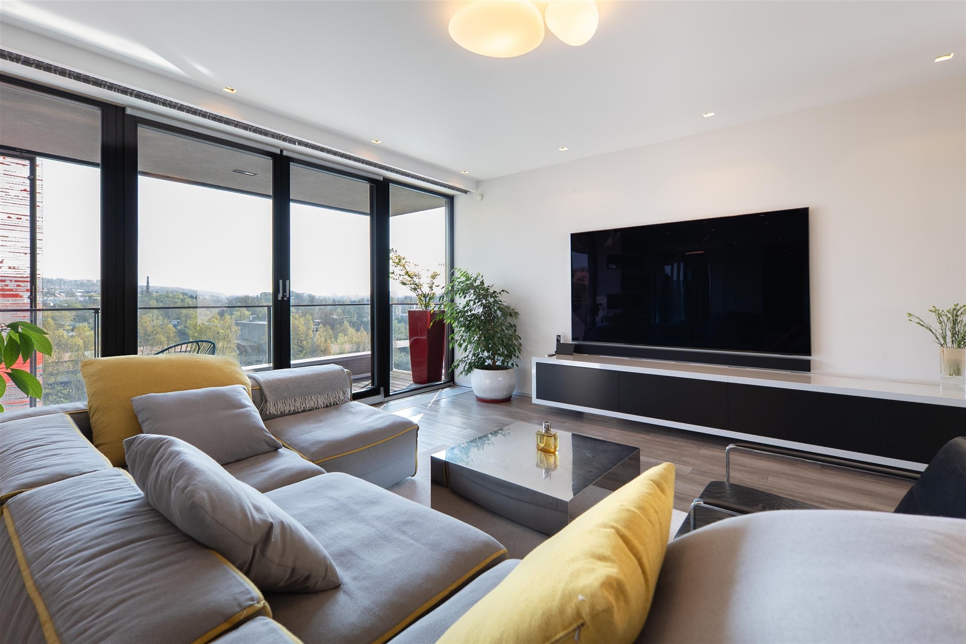 Světlý obývací pokoj s moderním designem, velká okna nabízejí panoramatický výhled, světlá parketová podlaha doplňuje elegantní interiér.
