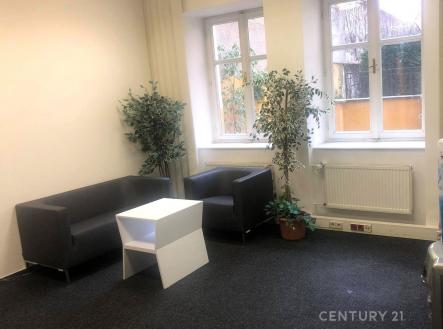 Světlý obývací pokoj s velkými okny a tmavým kobercem, moderní nábytek zahrnuje černou sedačku a křeslo, bílé stěny vytvářejí elegantní atmosféru. | Pronájem - kanceláře, 52 m²