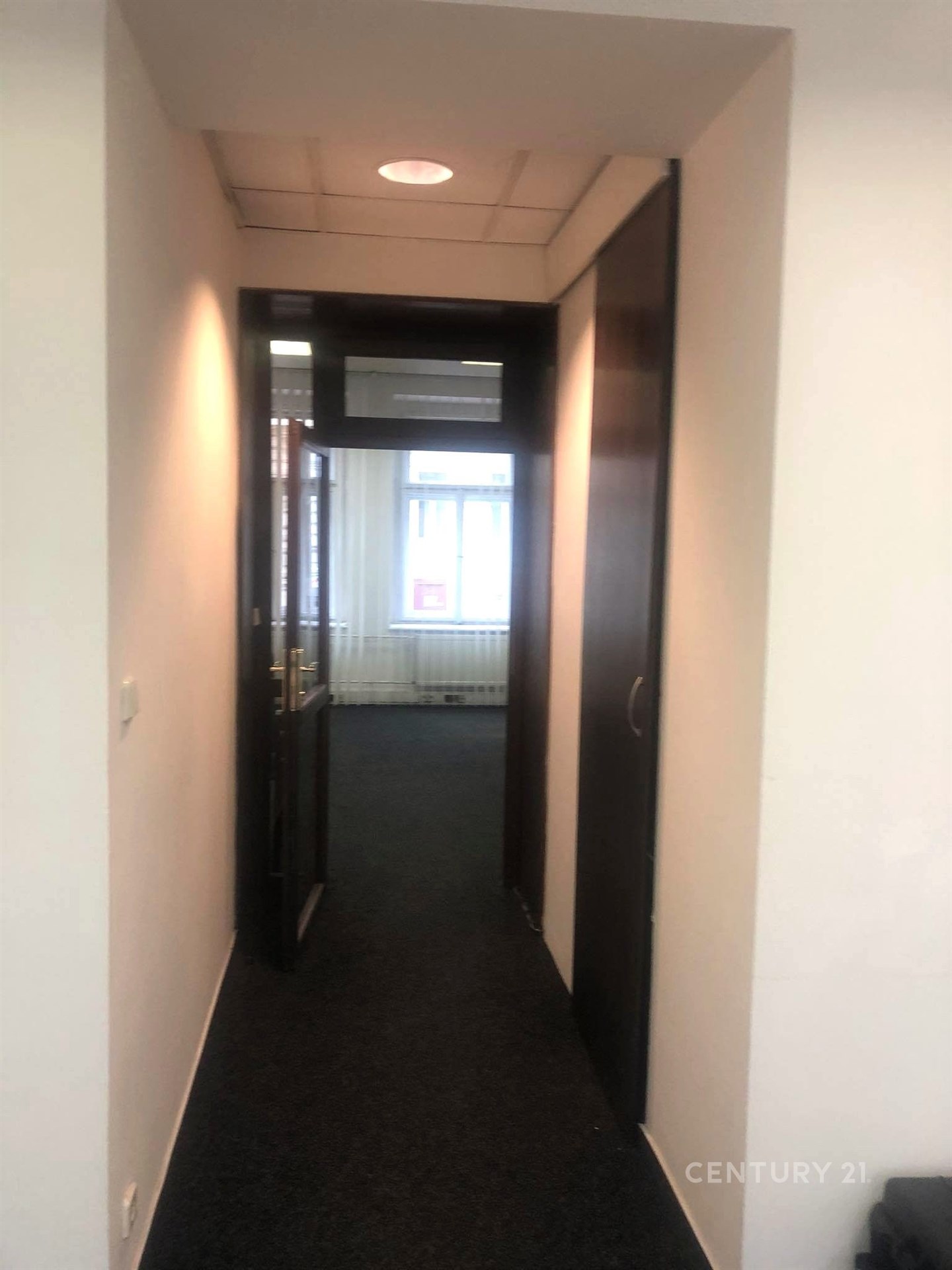 Úzká chodba s tmavým kobercem, moderní osvětlení, dřevěné dveře, vede do světlé místnosti s velkým oknem.