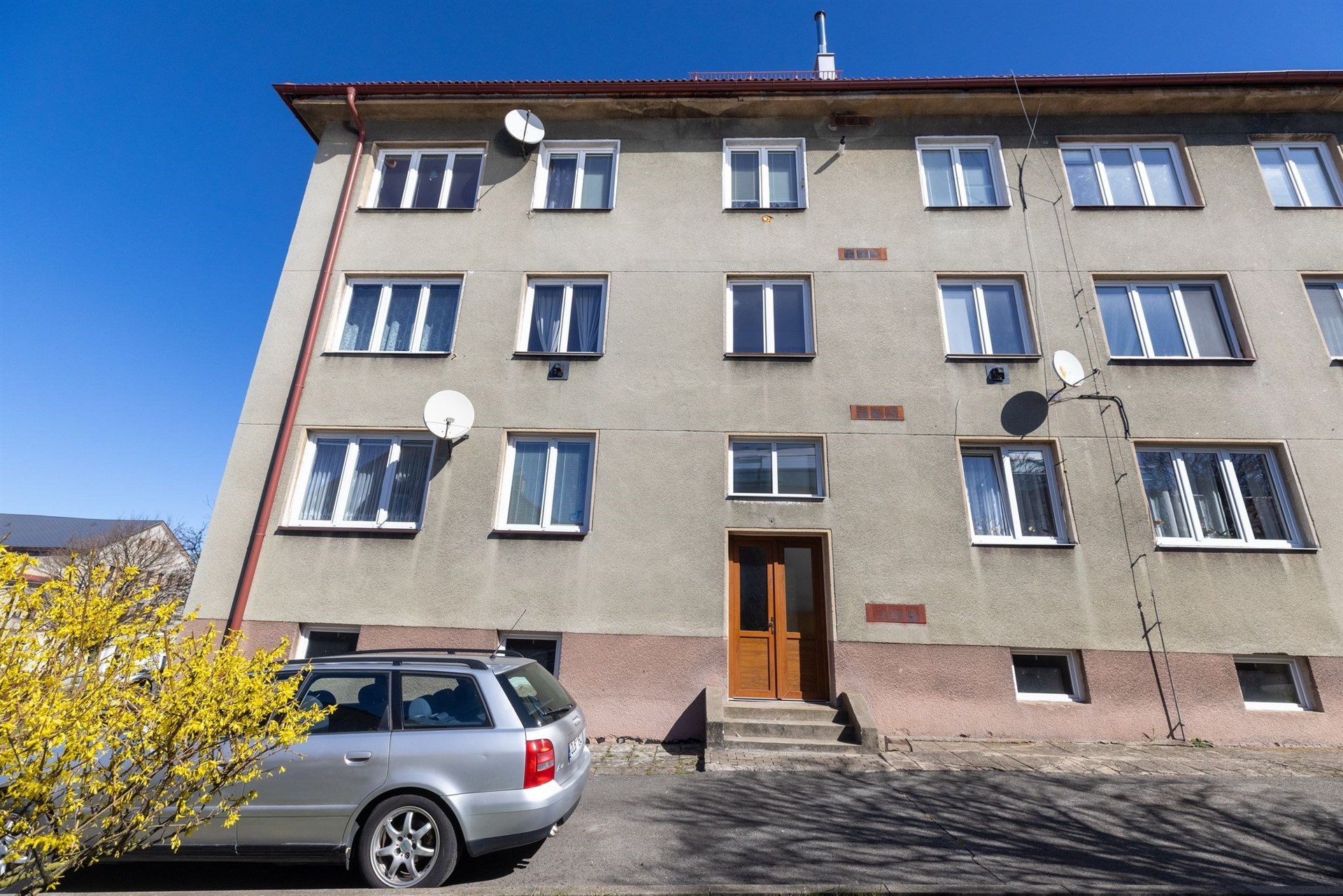 Čtyřpodlažní bytový dům s novými plastovými okny, fasádou v původním stavu a parkovištěm před budovou, ideální pro bydlení v klidné lokalitě.