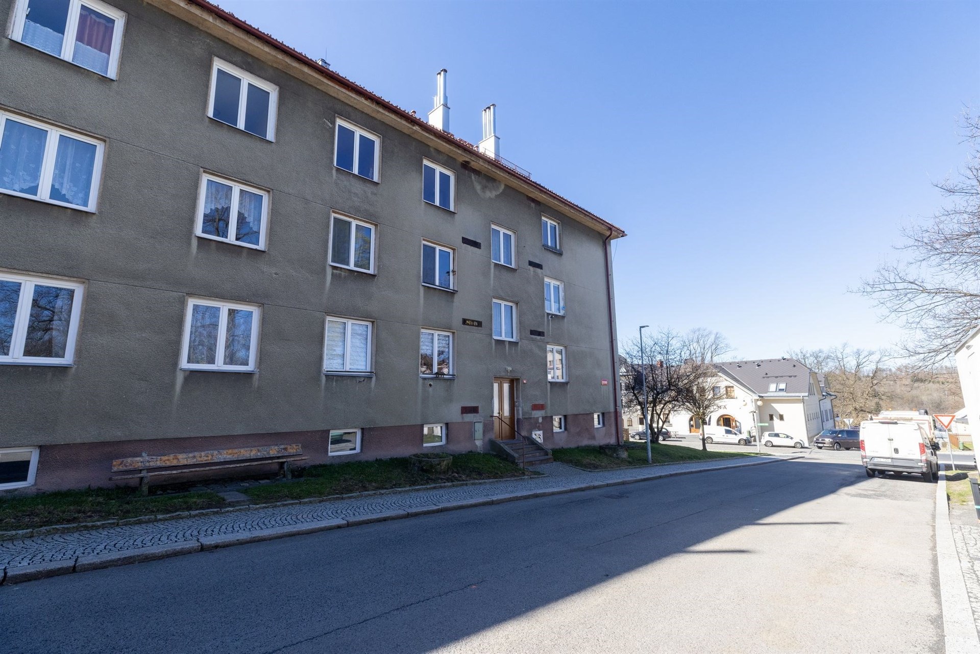 Čtyřpatrový bytový dům s šedou fasádou a bílými rámy oken v klidné ulici, vhodný pro investice nebo bydlení s výhledem na městské prostředí.