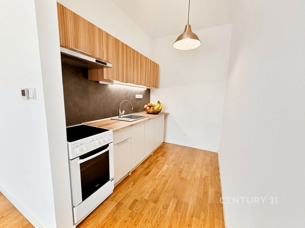 Moderní kuchyň s dřevěnou podlahou, světlými skříňkami a vestavěnými spotřebiči. Elegantní osvětlení nad pracovní deskou.