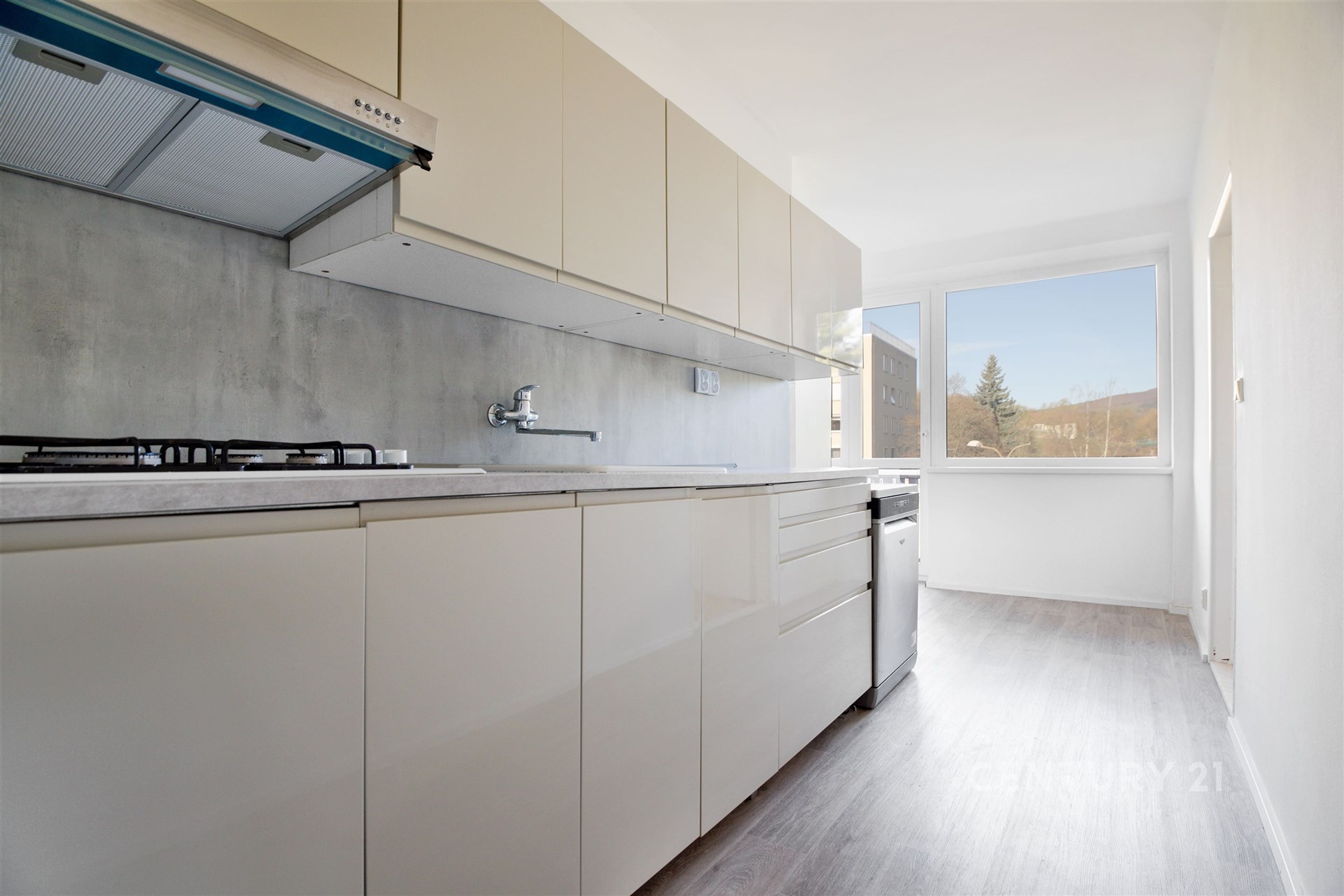 Světlá moderní kuchyň s vestavěnými skříněmi, parketová podlaha a velká okna zajišťující dostatek přirozeného světla.