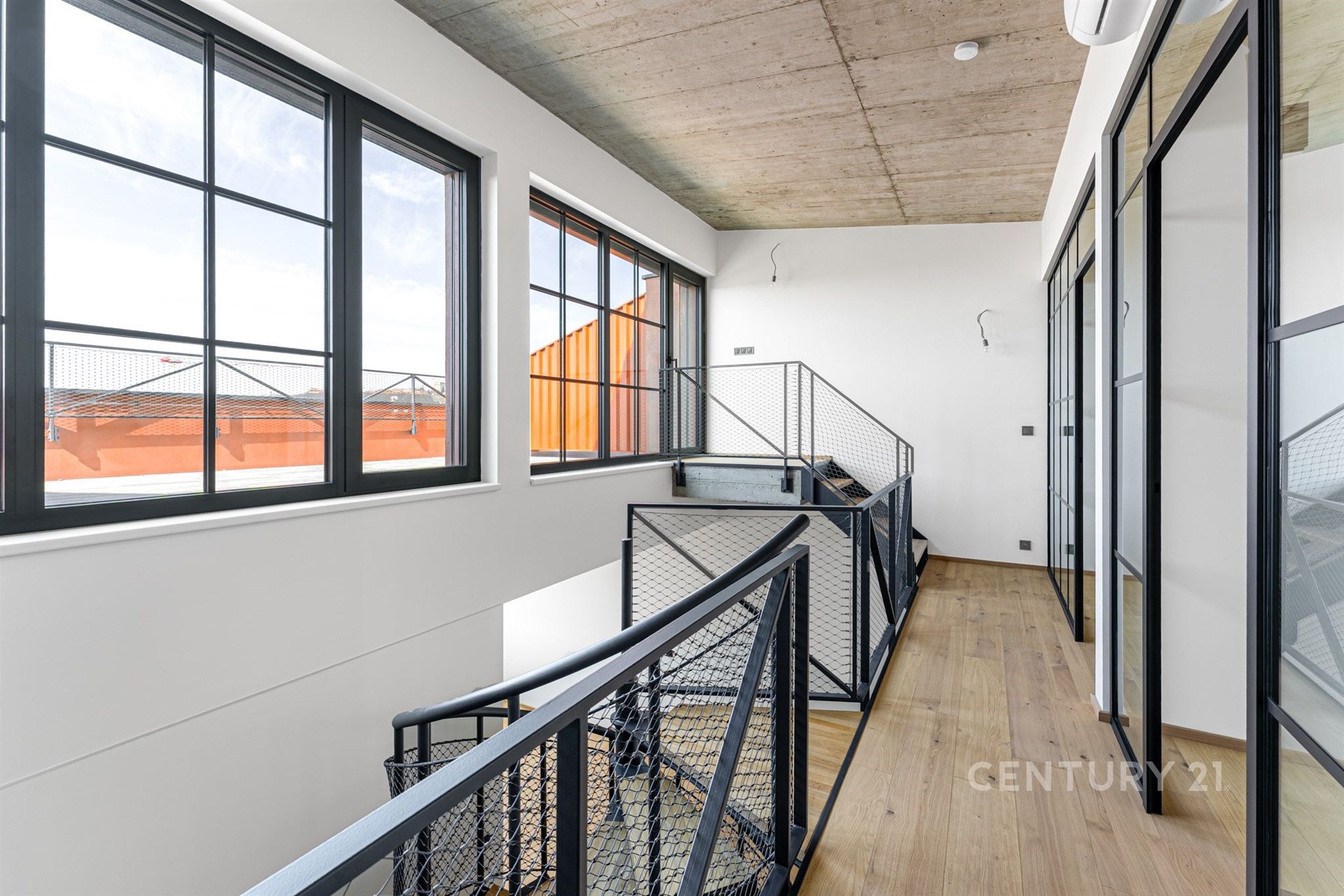 Světlá chodba s velkými okny, moderním průmyslovým stylem a dřevěnou podlahou. Skleněné stěny s kovovými rámy dodávají industriální vzhled.