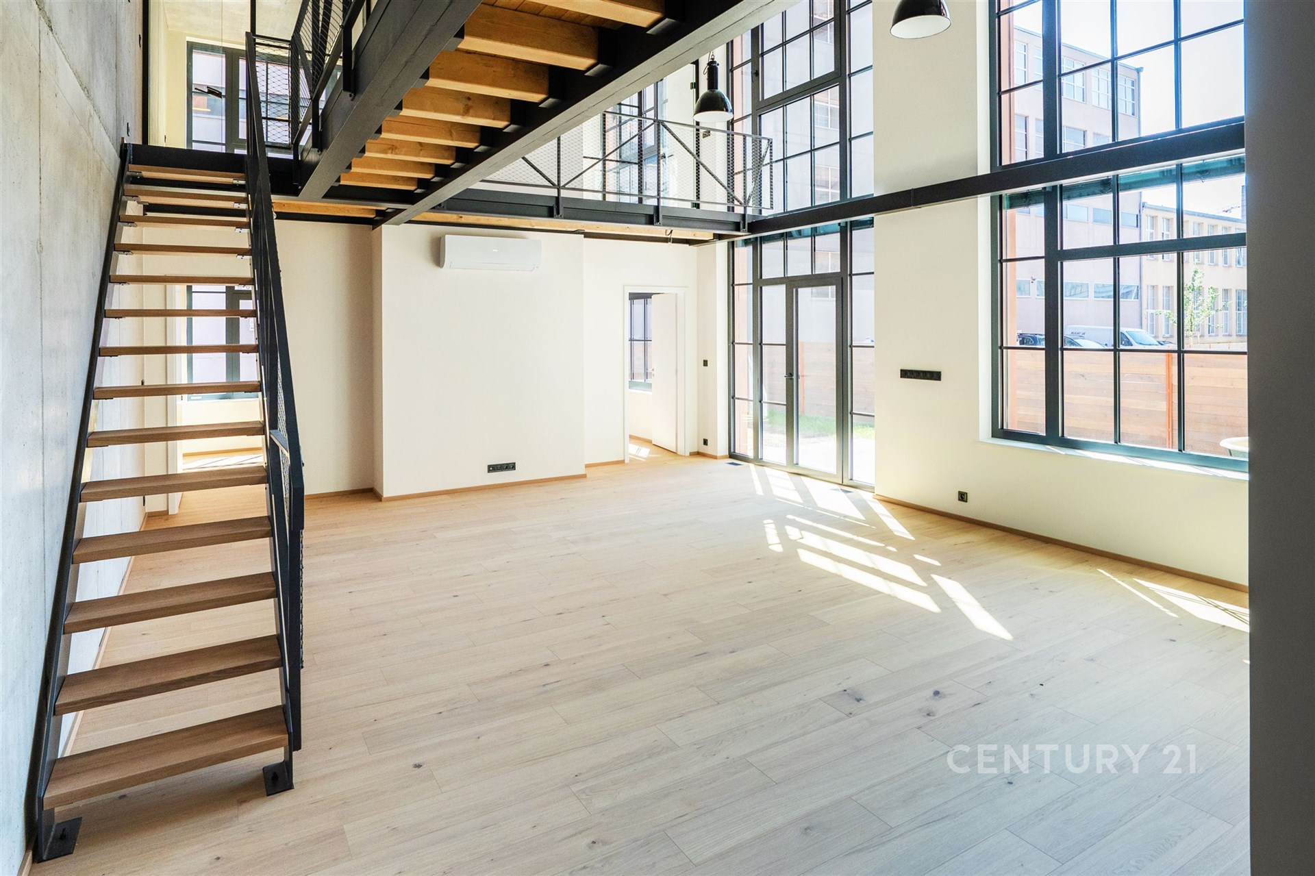 Světlý obývací pokoj s moderními velkými okny, parketovou podlahou a stylovým kovovým schodištěm, ideální pro vzdušný interiér.