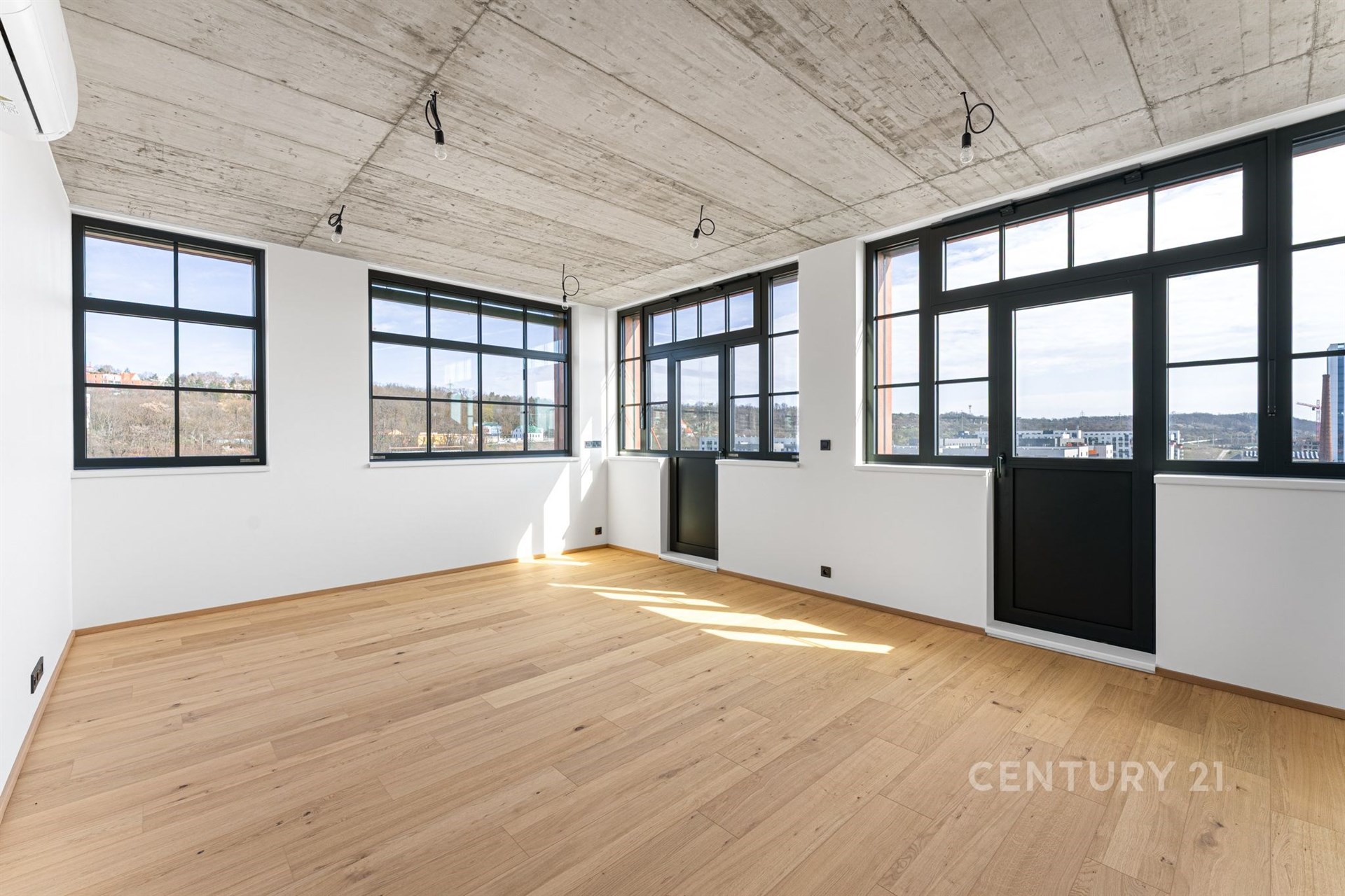 Světlý obývací pokoj s parketovou podlahou a velkými okny, moderní průmyslový styl s betonovým stropem a výhledem na město.