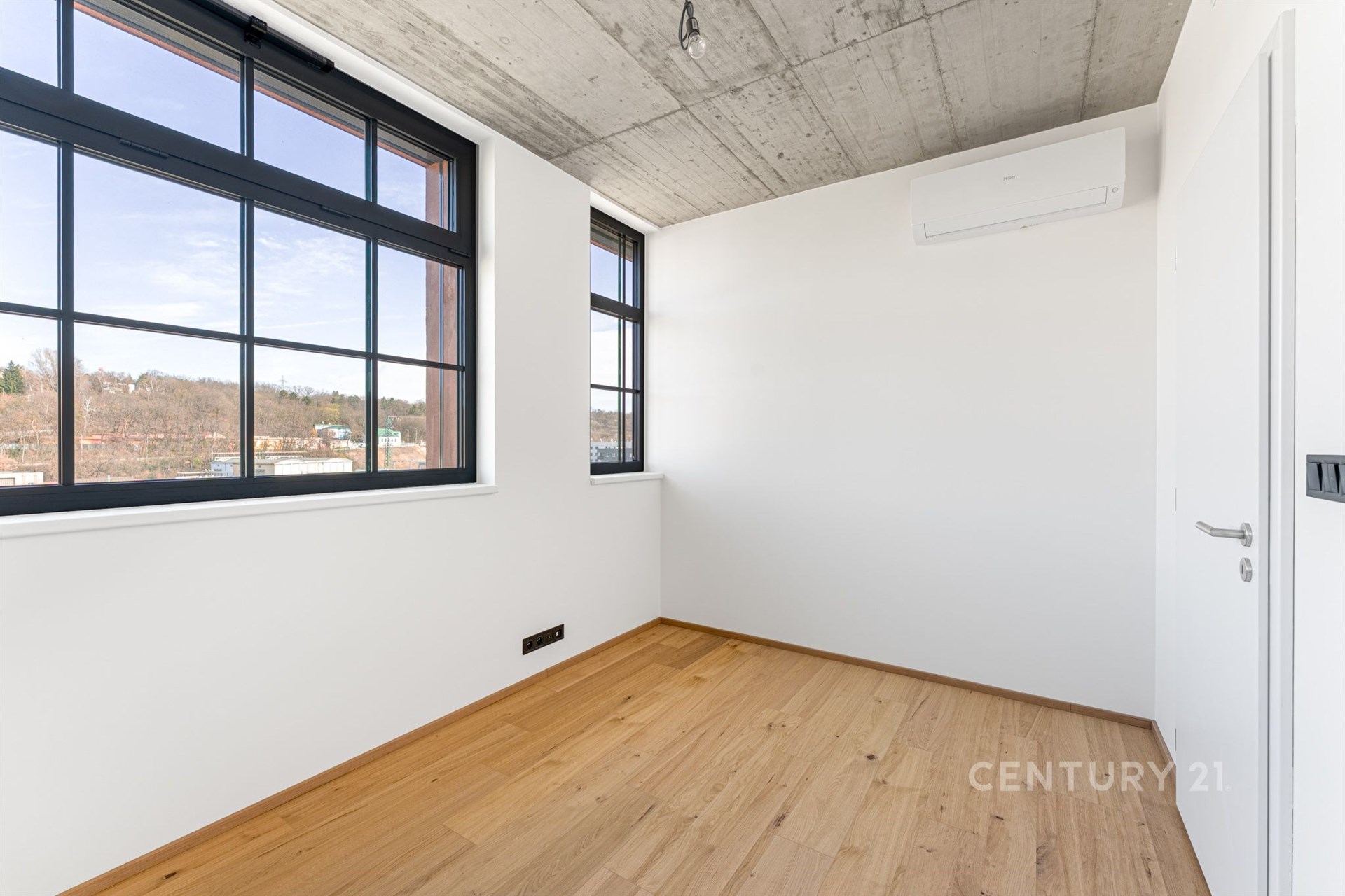 Světlý pokoj s velkými okny, moderní dřevěná podlaha a betonový strop, ideální pro rekonstruovaný industriální styl.