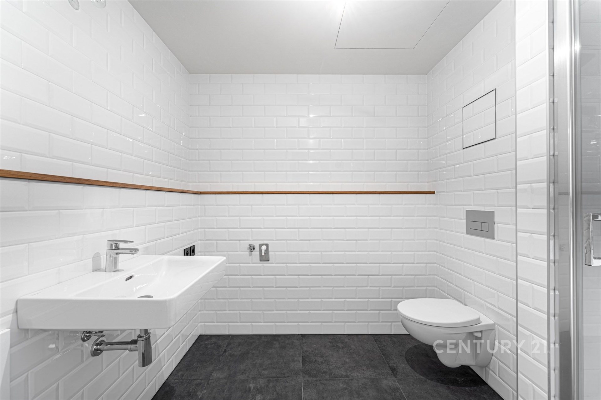 Moderní koupelna s bílými obklady, závěsnou toaletou a umyvadlem. Kombinace dlažby na podlaze a čistý design vytváří světlý prostor.