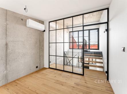 Průmyslově stylizovaná místnost s parketovou podlahou a betonovou stěnou, oddělena prosklenými dveřmi, poskytuje moderní a světelný interiér. | Prodej bytu, 2+kk, 49 m²