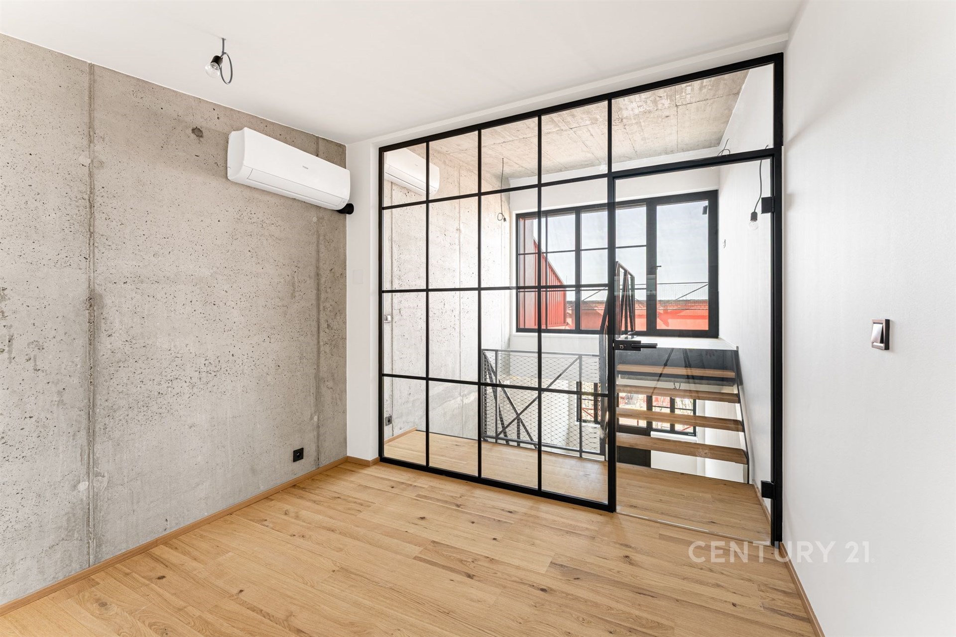 Moderní ložnice s parketovou podlahou, betonovou stěnou a velkými prosklenými dveřmi z černého rámu, které vedou k schodišti.