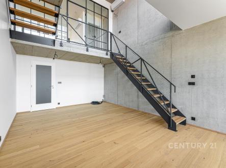 Světlý obývací pokoj s parketovou podlahou, moderní kovové schodiště, betonové stěny a velká okna poskytující přirozené světlo. | Prodej bytu, 2+kk, 65 m²