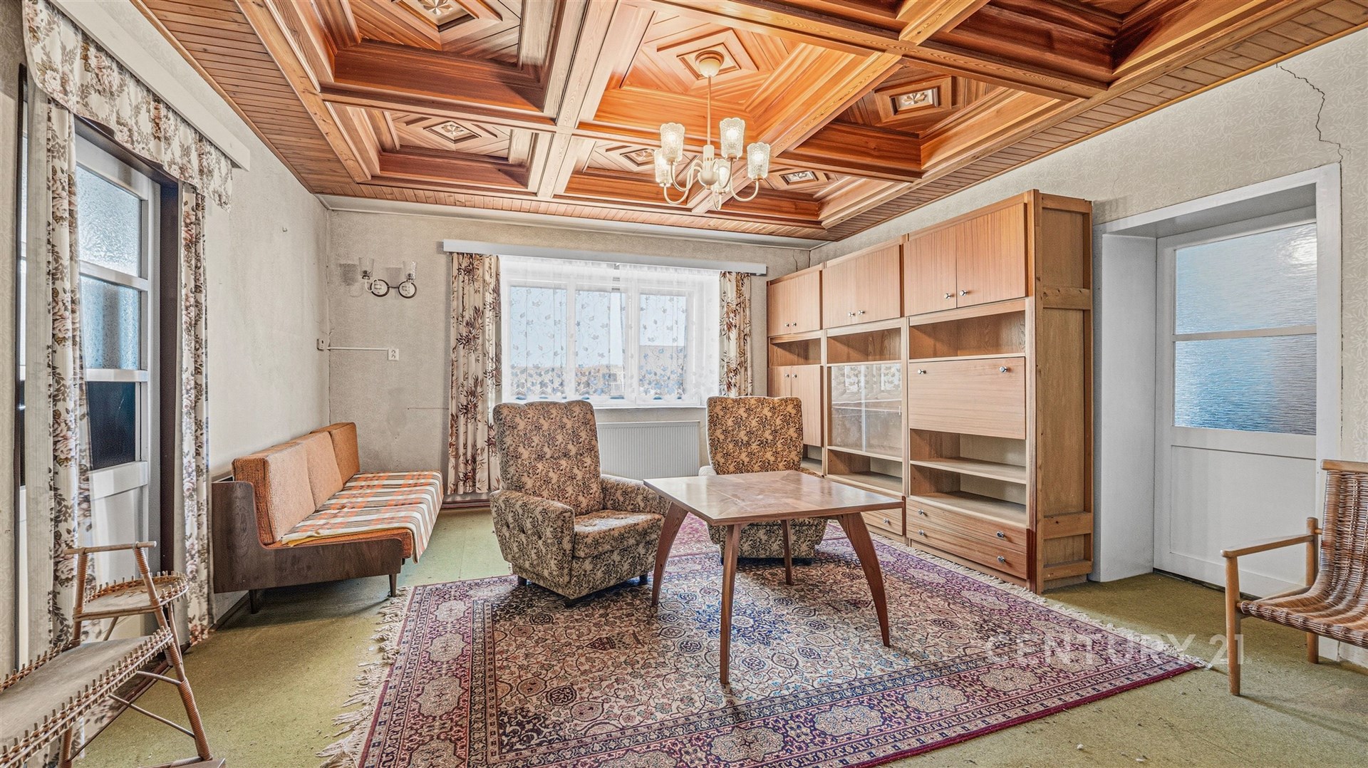 Obývací pokoj s dřevěným stropem, světlým kobercem a velkými okny, retro styl s nábytkem z 60. let, ideální pro renovaci.