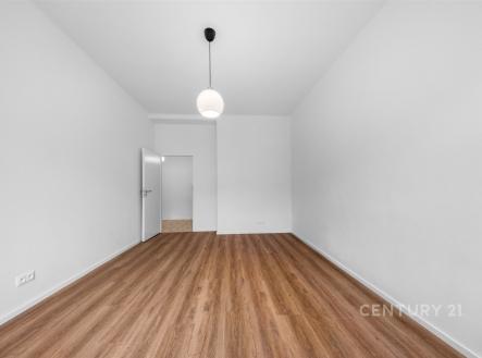 Prostorný pokoj s moderní dřevěnou podlahou, vysokým stropem a jednoduchým závěsným svítidlem, světlá výmalba a otevřené dveře do další místnosti. | Pronájem bytu, 2+kk, 52 m²