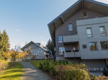 Moderní bytový komplex s šedou fasádou, parkovací plochou a klidným okolím, obklopený zelení a vzrostlými stromy, vhodný pro rodinné bydlení. | Prodej bytu, 1+kk, 32 m²