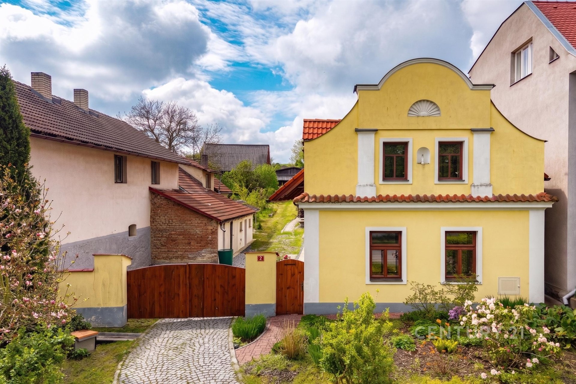 Tradiční fasáda rodinného domu s žlutou omítkou a dřevěnými okny, dlážděný chodník, udržovaná zahrada, historický styl.