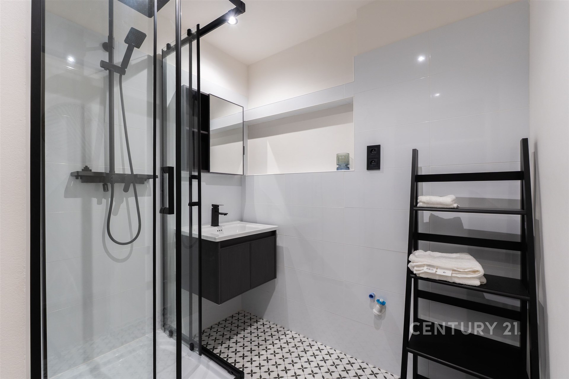 Moderní koupelna s proskleným sprchovým koutem, bílými dlaždicemi a dlažbou s geometrickým vzorem, elegantní černé prvky dodávají styl.