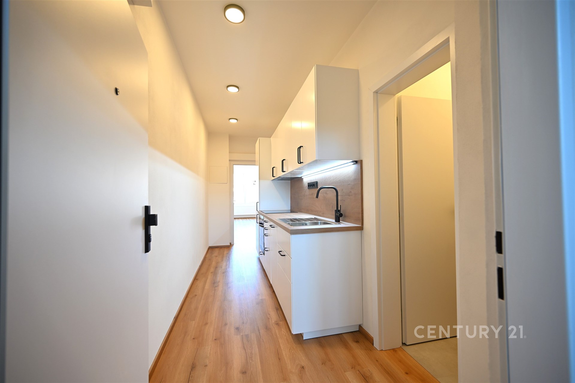 Úzká moderní kuchyň s parketovou podlahou, vestavěnými skříněmi a světlým osvětlením, ideální pro kompaktní byty.