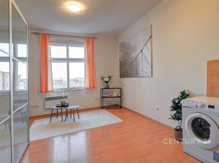 Prostorný obývací pokoj s laminátovou podlahou, velkým oknem a moderním dekorativním prvkem. Světlý interiér s oranžovými závěsy. | Prodej bytu, garsoniéra, 26 m²