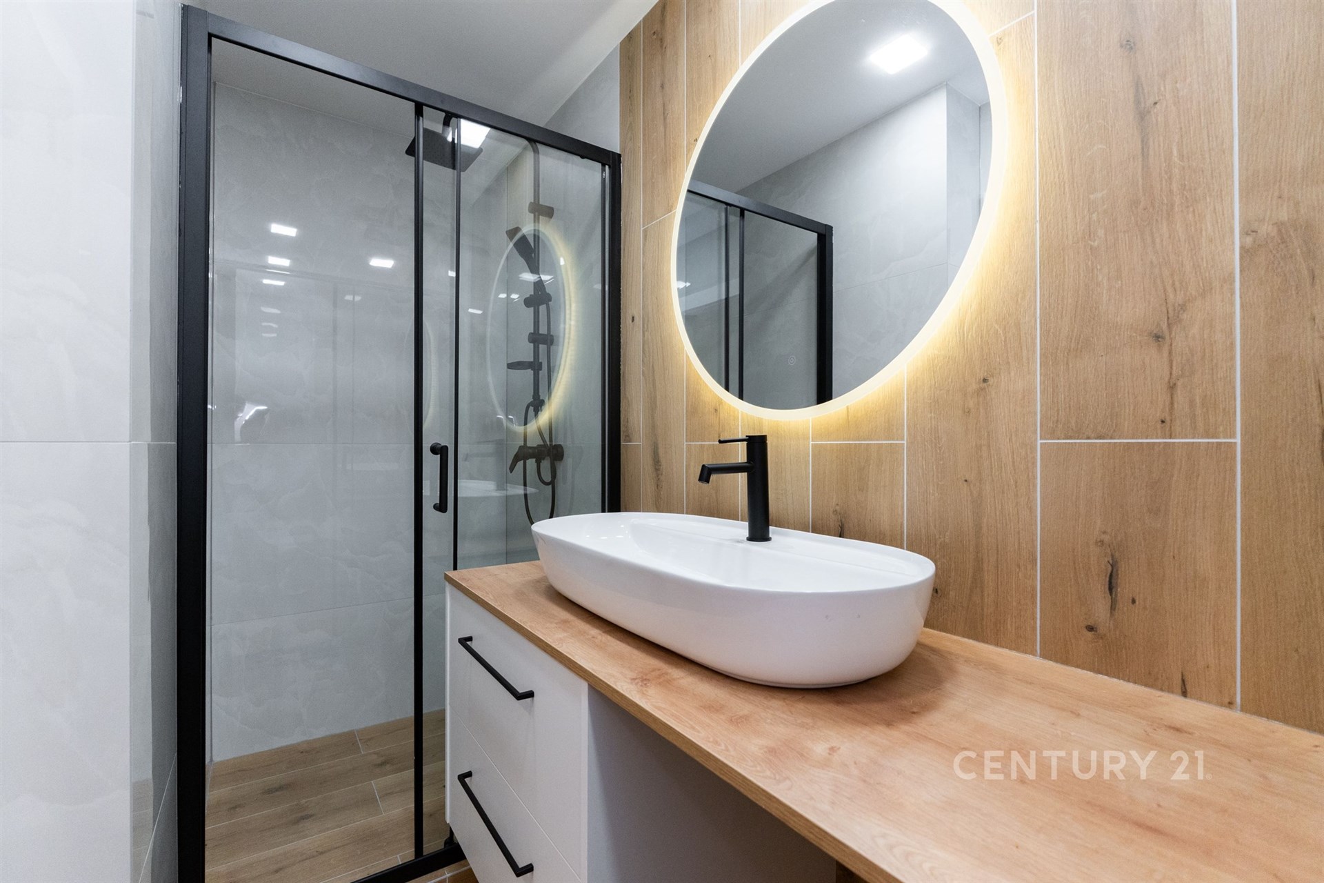 Moderní koupelna s velkým zrcadlem, dřevěným obkladem a sprchovým koutem. Stěny z kvalitní dlažby dodávají elegantní vzhled.