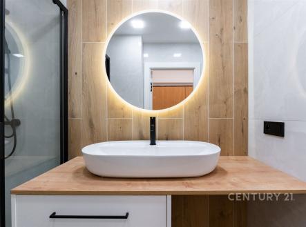 Moderní koupelna s dřevěným obkladem, kulaté zrcadlo s LED osvětlením a umyvadlo s černou baterií, vytvářející elegantní vzhled. | Prodej bytu, 2+kk, 35 m²
