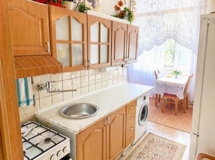 Kuchyň s dřevěnými skříněmi, bílou dlažbou a velkým oknem, které poskytuje dostatek světla, součástí je i malý jídelní kout. | Prodej bytu, 2+1, 57 m²