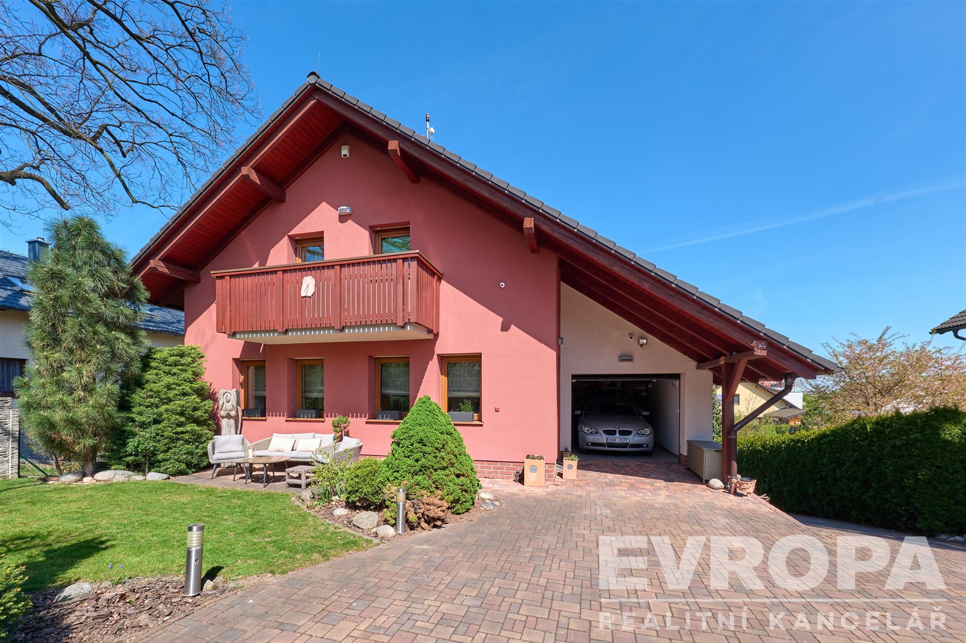 Růžový rodinný dům s prostornou garáží, dlážděný příjezd a udržovaná zahrada, dřevěný balkon a moderní vzhled pro pohodlné bydlení.
