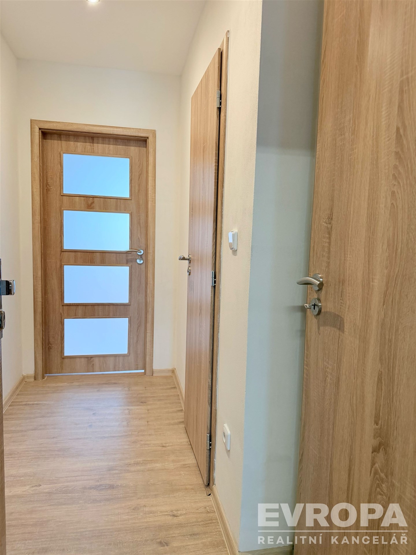 Úzká chodba s dřevěnou podlahou a moderními vnitřními dveřmi, světlé stěny a elegantní design, ideální pro rodinný byt.
