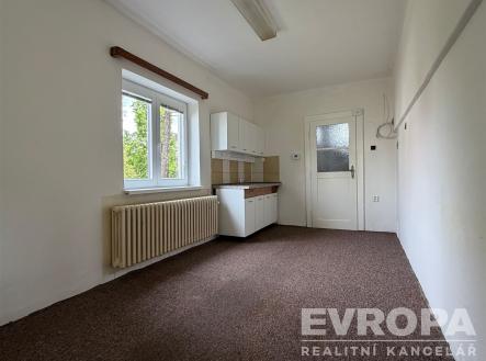 Kuchyňský kout s kobercem, bílými skříňkami, světlým interiérem a velkým oknem. Prostor pro renovaci, základní vybavení kuchyně. | Pronájem bytu, 1+kk, 27 m²