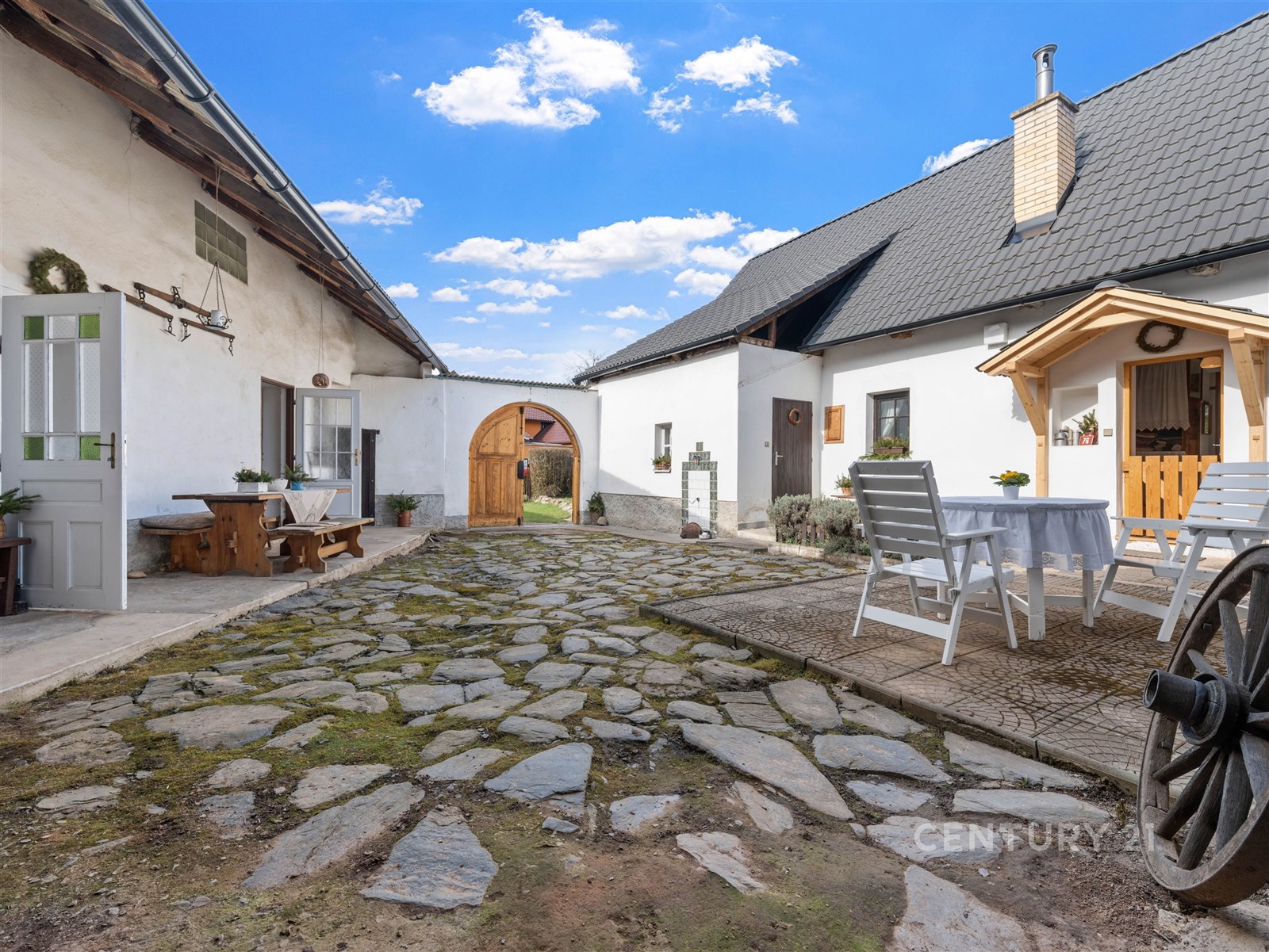 Venkovní dvůr s kamennou dlažbou a dřevěným nábytkem, rustikální styl, bílá fasáda budov, ideální pro relaxaci.