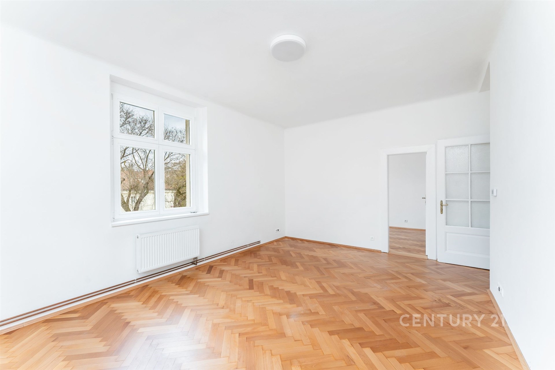 Světlý obývací pokoj s parketovou podlahou, velká okna a bílými stěnami, moderní styl a prostorný interiér vhodný pro rodinné bydlení.