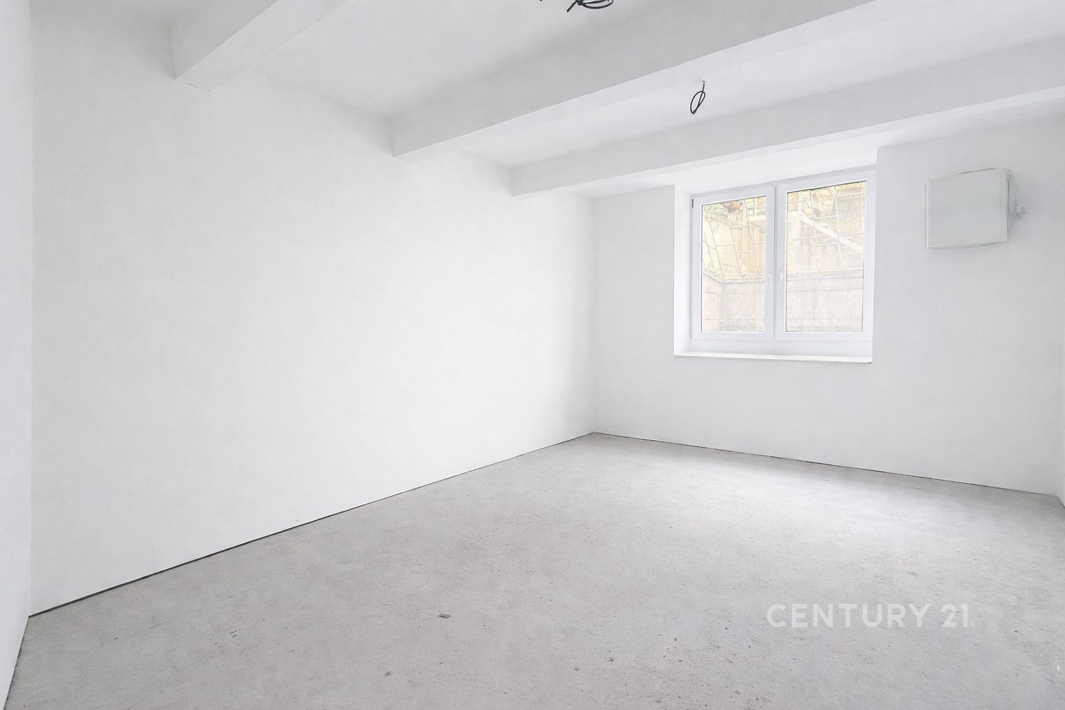 Rekonstruovaný světlý pokoj s bílými stěnami, betonovou podlahou a velkým oknem poskytujícím přirozené světlo.