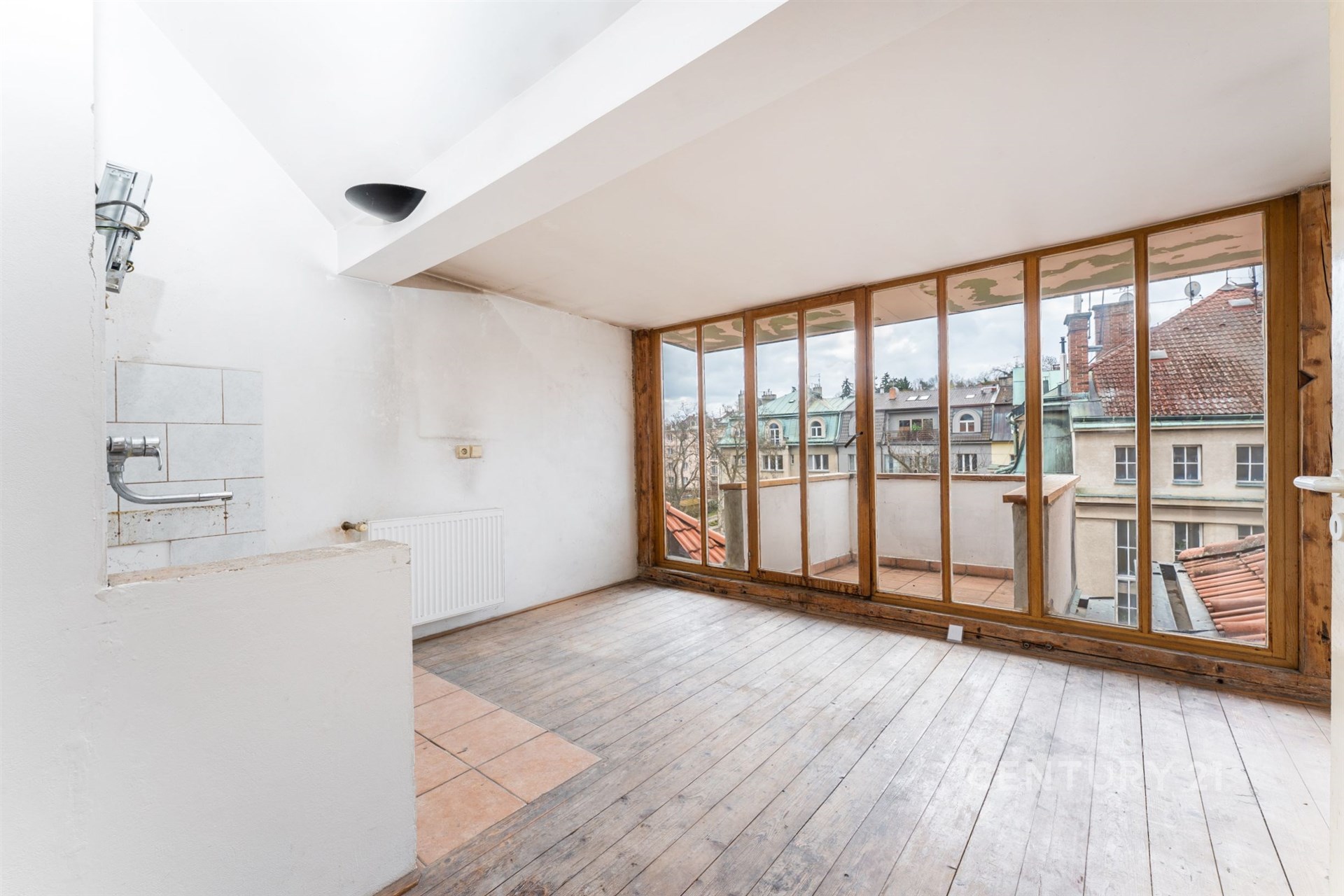Podkrovní místnost s dřevěnými podlahami a velkými okny, nabízející výhled na historické budovy, ideální pro moderní rekonstrukci.