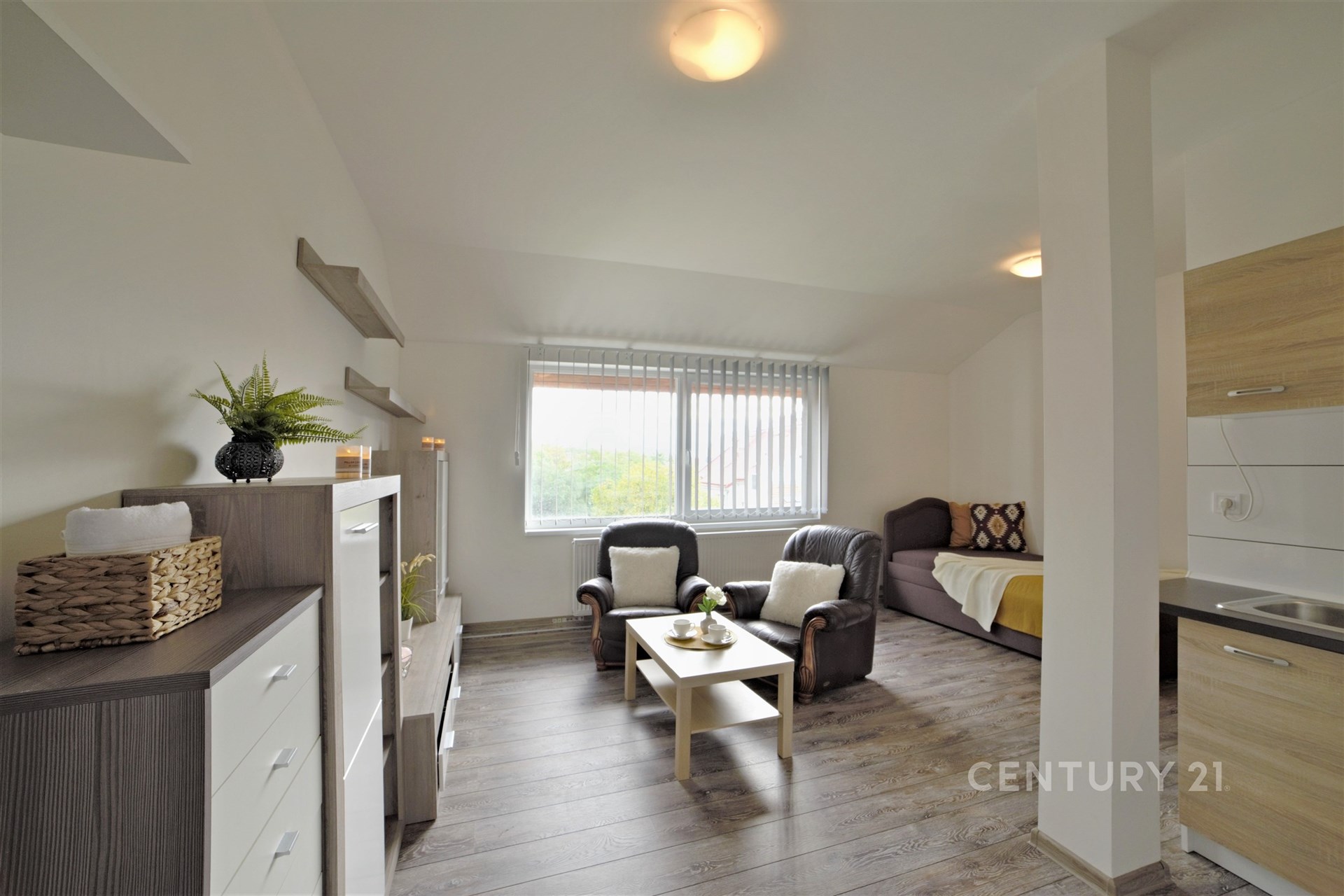 Světlý obývací pokoj s parketovou podlahou a velkými okny, moderní interiér s elegantním nábytkem a dekoracemi, propojený s kuchyňským koutem.