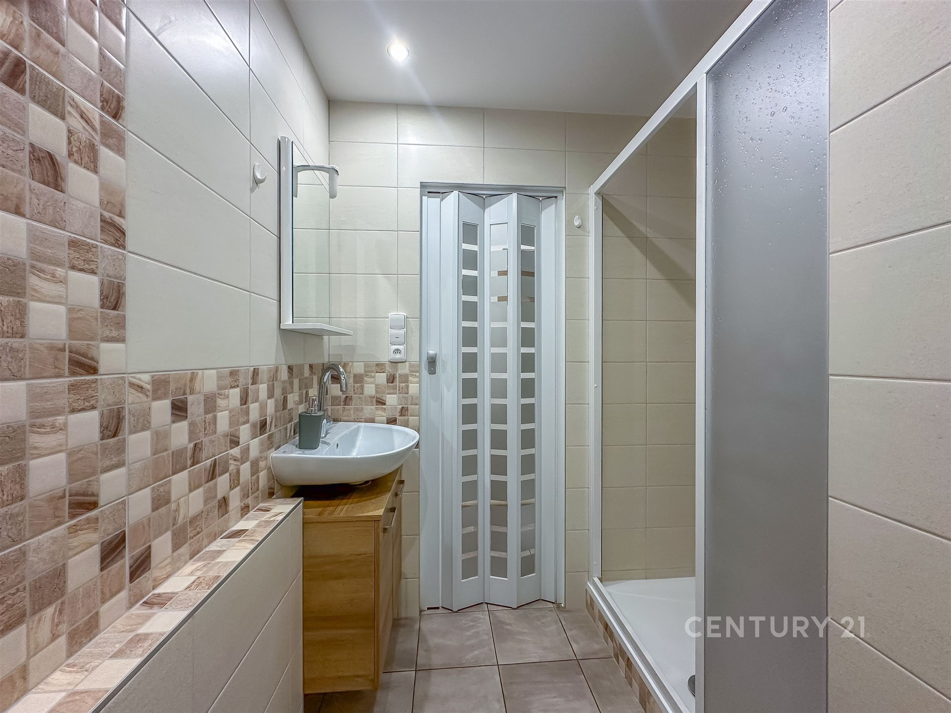 Koupelna s moderní dlažbou, sprchovým koutem a dřevěnou skříňkou, světlé dlaždice vytvářejí čistý a prostorný vzhled.