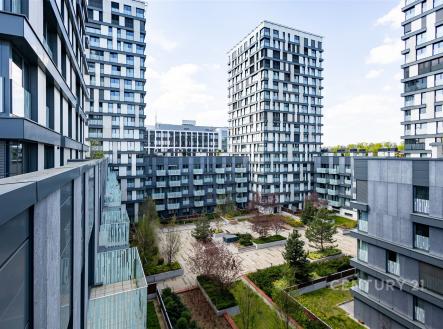 Výhled z balkonu na moderní bytový komplex s upraveným vnitroblokem, velkými okny a šedou fasádou. | Pronájem bytu, 1+kk, 37 m²