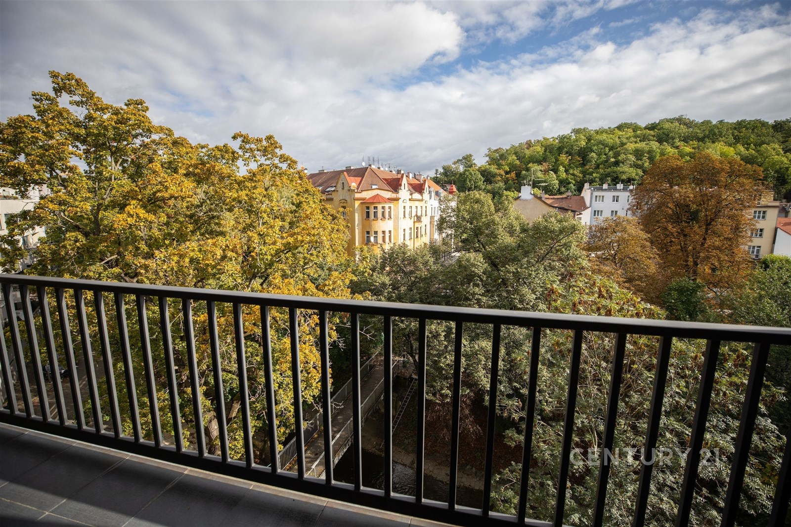 Výhled z balkonu s moderním zábradlím, nabízí panoramatický pohled na zelené stromy a historické budovy, ideální pro relaxaci ve městě.