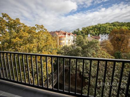 Výhled z balkonu s moderním zábradlím, nabízí panoramatický pohled na zelené stromy a historické budovy, ideální pro relaxaci ve městě. | Prodej bytu, 3+kk, 79 m²