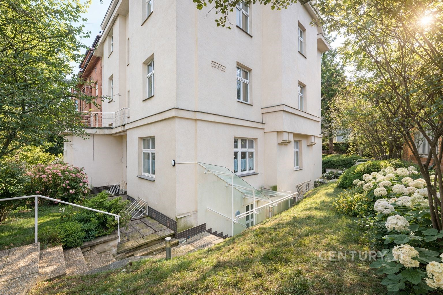 Rohový exteriér bytového domu s květinovou zahradou, bílá fasáda, velká okna, okolní zeleň vytváří klidné a příjemné prostředí.