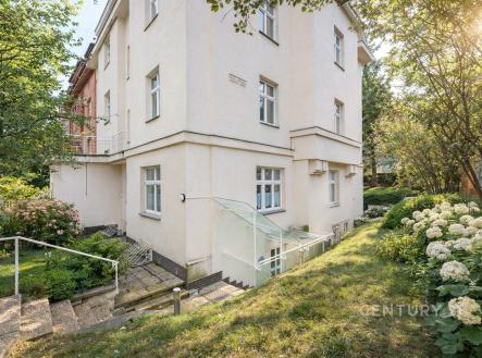 Rohový exteriér bytového domu s květinovou zahradou, bílá fasáda, velká okna, okolní zeleň vytváří klidné a příjemné prostředí. | Prodej bytu, 3+kk, 75 m²
