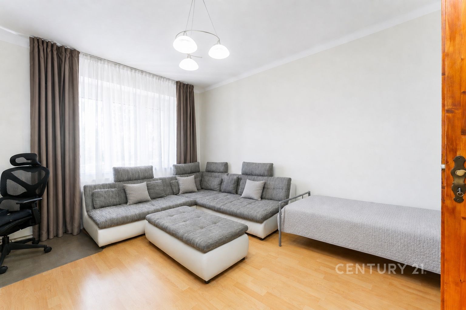 Světlý obývací pokoj s parketovou podlahou, moderní šedá sedací souprava, velká okna s bílými závěsy, minimalistický styl interiéru.