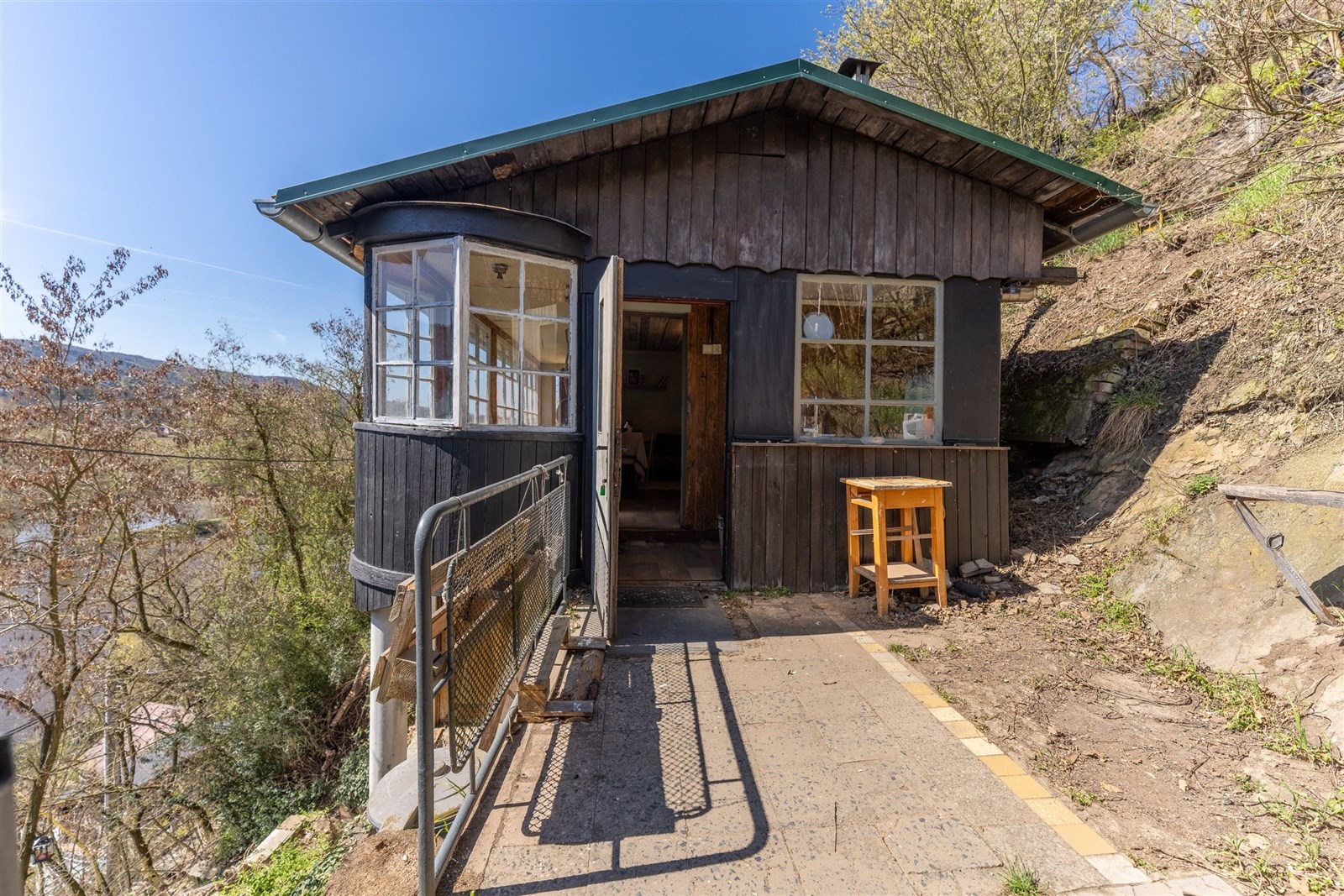 Dřevěná chata s verandou a velkými okny, umístěná na svažitém terénu. Přírodní materiály a rustikální vzhled dodávají autentický charakter.