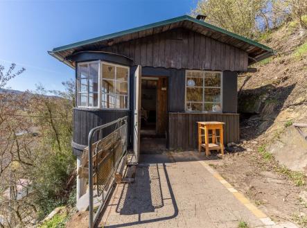 Dřevěná chata s verandou a velkými okny, umístěná na svažitém terénu. Přírodní materiály a rustikální vzhled dodávají autentický charakter. | Prodej - dům/vila, 47 m²