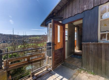 Venkovní terasa s dřevěnou podlahou a výhledem na krajinu, ideální pro relaxaci, útulný styl, původní dřevěné obložení stavby. | Prodej - dům/vila, 47 m²