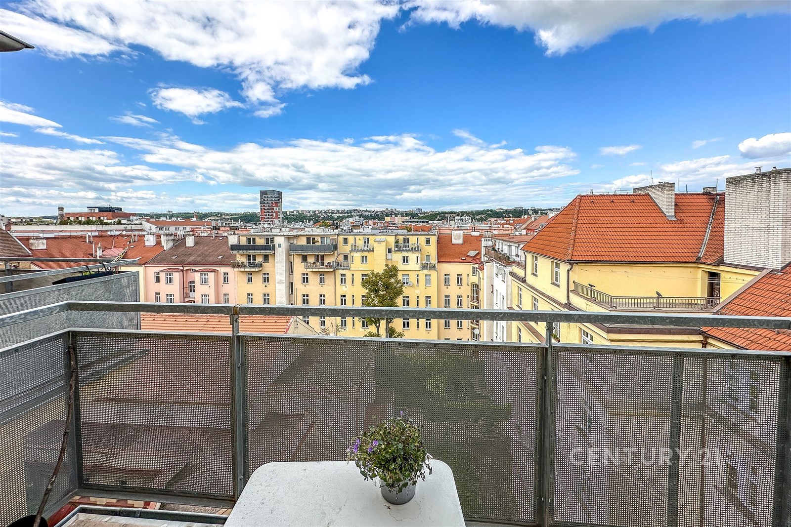 Balkon s kovovým zábradlím a výhledem na město, světlé cihlové budovy s červenými střechami, modrá obloha s mraky