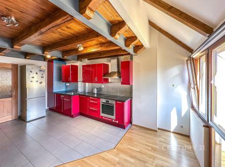 Moderní kuchyň s červenými lesklými skříňkami, nerezovými spotřebiči a dlažbou, propojená se světlým obývacím prostorem s parketovou podlahou. | Pronájem bytu, 4+kk, 102 m²